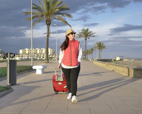 Jeune voyageuse se promène le long de la promenade au bord de la mer avec une valise rouge, vue de face — Photo