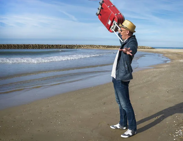 Un jeune homme positif se réjouit debout au bord de la mer, lève une valise rouge — Photo