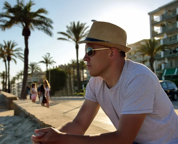 Jeune homme attrayant dans un chapeau et des lunettes de soleil est assis sur le front de mer par la mer — Photo