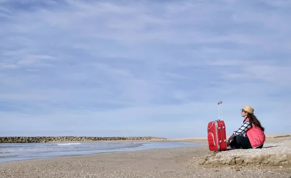 Jolie fille de voyage brune assise avec une valise rouge au bord de la mer, contre le ciel bleu, endroit pour le texte — Photo