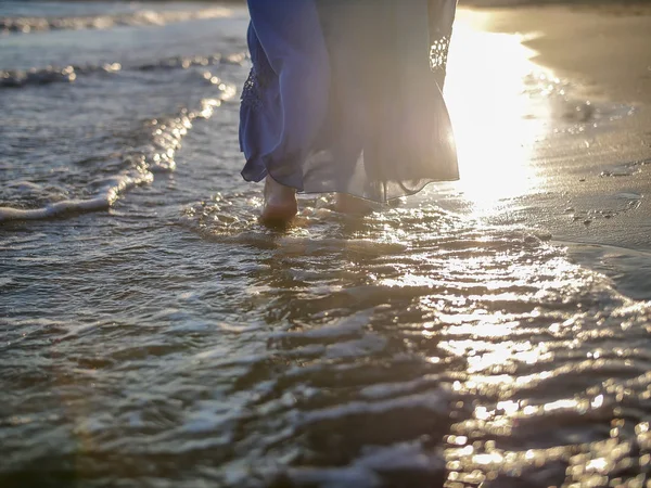 Une fille dans une longue jupe bleue marche le long de la plage, l'éblouissement du soleil sur les vagues, une fille va sur le côté du coucher du soleil — Photo