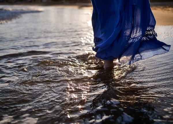 A young woman in a blue long skirt walks along the beach, the glare of the sun on the waves, sunset — Stock Photo, Image