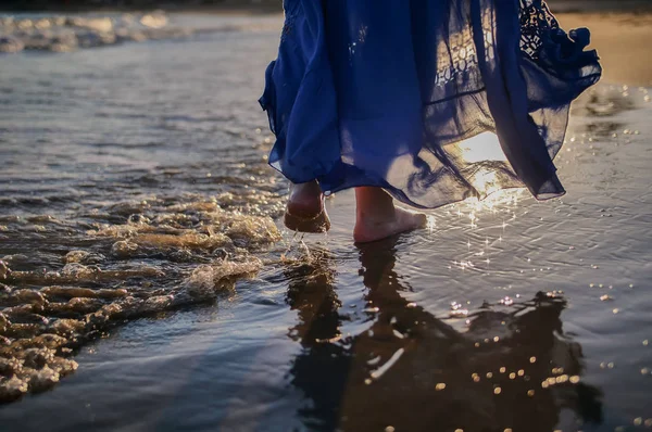 Ben af en ung kvinde i en blå lang nederdel går langs stranden, solen blænding på bølgerne, solnedgang - Stock-foto