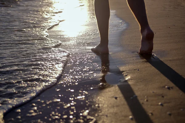 Slanke ben af en mand, der går langs stranden, skummende bølger vasker sandet, solnedgang på stranden - Stock-foto