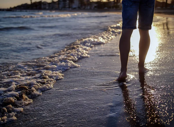 Un homme en jeans se promène le long de la plage, les vagues de mousse lavent le sable, coucher de soleil sur la plage — Photo