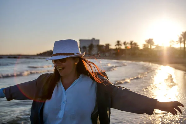 Jolie jeune femme dans un chapeau blanc et veste bleue tourne et s'amuse sur la plage, au coucher du soleil — Photo