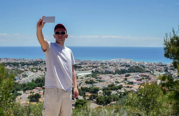 Um jovem fala num telemóvel, faz videochamadas enquanto está numa montanha junto ao mar Imagens Royalty-Free