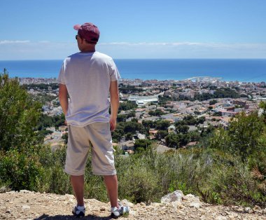 Genç adam turist bir dağ üzerinde duruyor ve dağın eteklerinde denize yakın şehir bakar