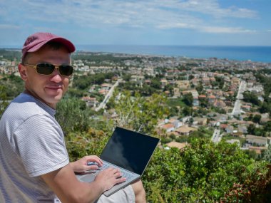 Serbest çalışan bir adam kameraya bakar ve altında güzel bir manzara, deniz tarafından bir şehir ile, bir dağ üzerinde oturur