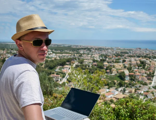 Un homme indépendant en chapeau regarde la caméra assis sur une montagne, avec un beau paysage, une ville au bord de la mer — Photo