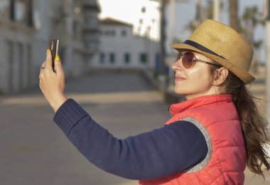 Şapka lı ve gözlüklü çekici genç kadın güneşli bir sonbahar gününde selfie çekiyor