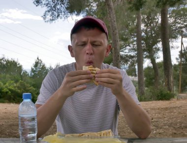 Piknikte genç çekici adam shawarma yeme ahşap bir masada oturan, doğada yemek, fast food