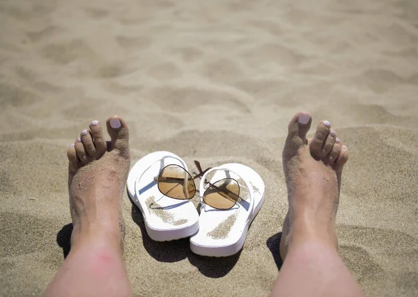 Tøfler og solbriller ved siden af kvindelige ben på stranden, afslapningskoncept - Stock-foto