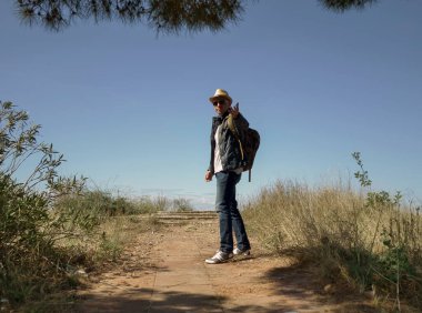 Şapkalı ve sırt çantalı bir adam bir tepenin gözlem güvertesinde duruyor ve elini uzatıyor.