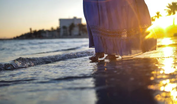 En pige i en blå lang nederdel går alene langs stranden under en smuk solnedgang - Stock-foto