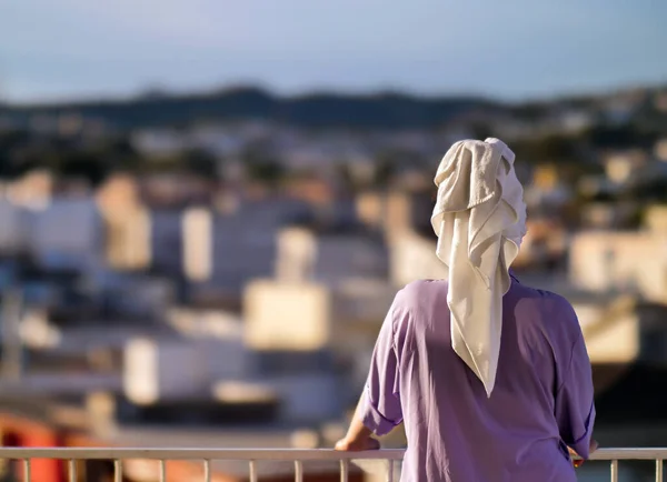 Une femme se tient sur le balcon avec une serviette blanche sur la tête sur le fond de la ville, vue arrière — Photo