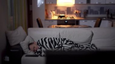 Hasta bir adam akşam kanepede yatar. TV 'nin yanında, öksürür, kamera hareket eder.