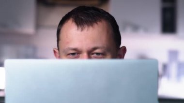 Gri gözlü yakışıklı adam laptop ekranının arkasında çalışıyor, yakın plan, kamera hareketi.