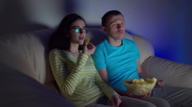 Akşam geç saatlerde genç bir çift TV 'de film izleyip kanepede otururken cips yiyor.