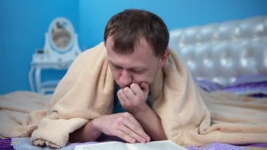 Bir adam yatağa uzanıp kitap okuyor, sonra başını battaniyeyle örtüyor ve uykuya dalıyor.