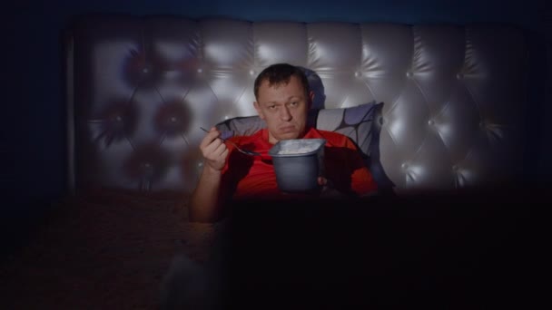疲惫的男人晚上躺在电视旁边的床上 手里拿着一桶冰淇淋 镜头在移动 — 图库视频影像