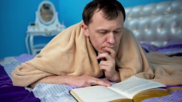 Attraente giovane uomo che legge un libro mentre si trova su un letto in una camera da letto — Video Stock