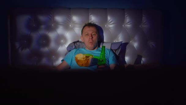 Un hombre pasa la noche en el televisor con una botella de cerveza y un tazón de papas fritas — Vídeo de stock