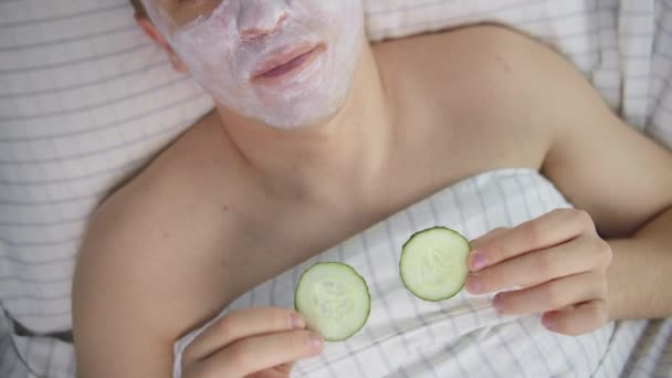 Jovem homem atraente com uma máscara cosmética em seu rosto e fatias de pepino em seus olhos, vista superior, movimento da câmera — Vídeo de Stock