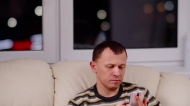 Elinde telefon olan genç bir adam pencerenin önünde oturur, kamera yukarı hareket eder.