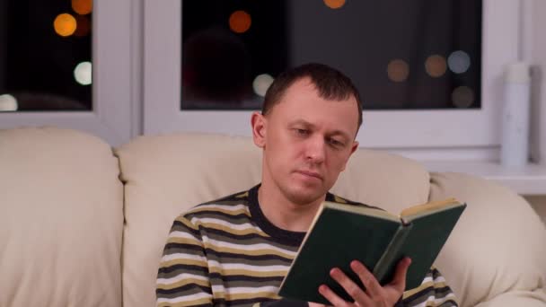 Attraktiv ung man sitter på en soffa med en bok, mot bakgrund av ett fönster — Stockvideo