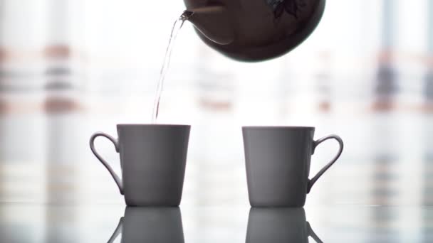 Zwei weiße Keramikbecher auf hellem Fensterhintergrund, aus einer alten Metall-Teekanne kochendes Wasser in Becher gießen — Stockvideo