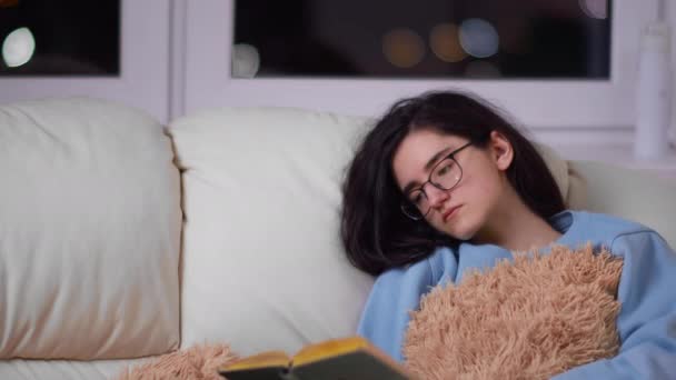 Cute młoda brunetka dziewczyna z okularami w godzinach wieczornych na kanapie czyta książkę, przestrzeń kopiowania — Wideo stockowe