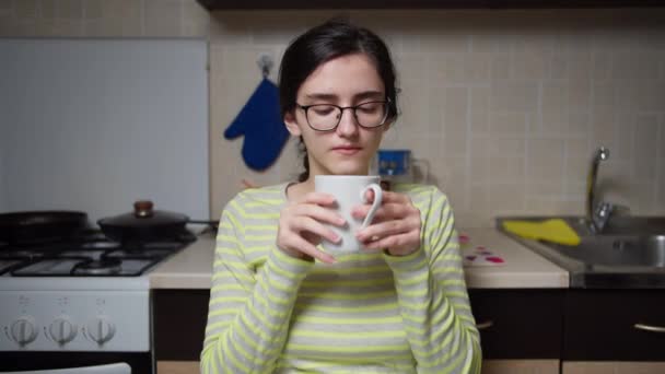 Flicka i glas sitter i köket, dricker ur en vit mugg och tittar på kameran — Stockvideo