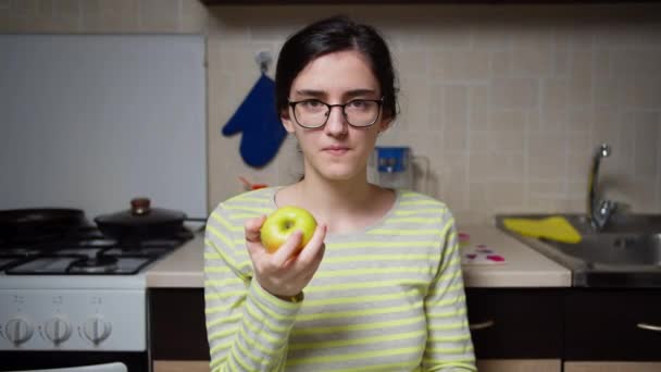Το κορίτσι με τα γυαλιά κάθεται στο τραπέζι της κουζίνας και τρώει ένα μήλο, κοιτάζει την κάμερα. — Αρχείο Βίντεο