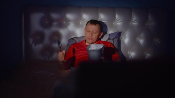 一个年轻人笑着躺在床上 手里拿着一桶冰激凌 躺在电视机前 摄像机在晃动 — 图库视频影像