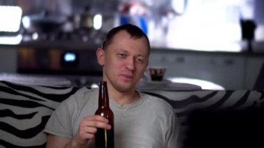Akşamları genç ve mutlu bir adam kanepede oturur, televizyon izler ve arka planda bira içer.