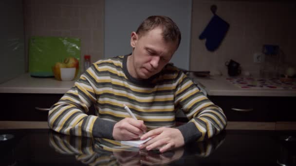 若い男がノートにメモを取り台所の夕方に座って — ストック動画