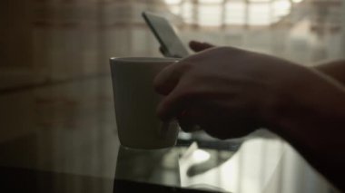 Cep telefonu olan bir adam bir fincan sıcak kahveyle masada oturuyor, pencerenin arka planında, yakın plan.