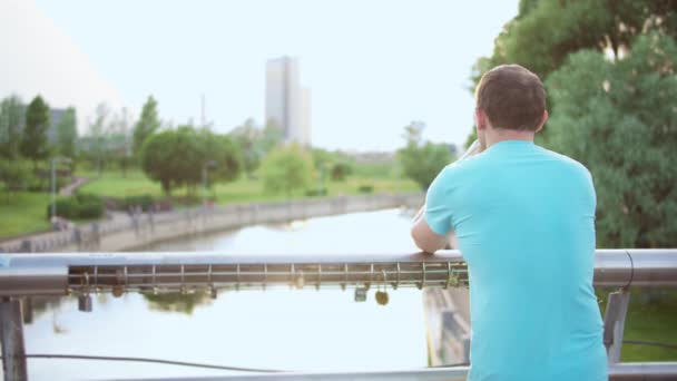 Man Står Park Bro Och Tittar Floden Kamerarörelse Bakre Kopieringsutrymme — Stockvideo