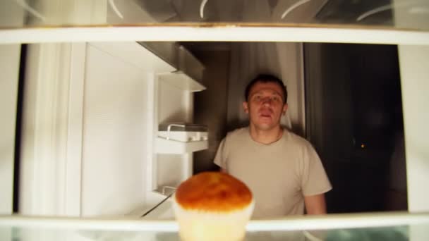 De man opent de koelkast en neemt een cupcake van de plank — Stockvideo