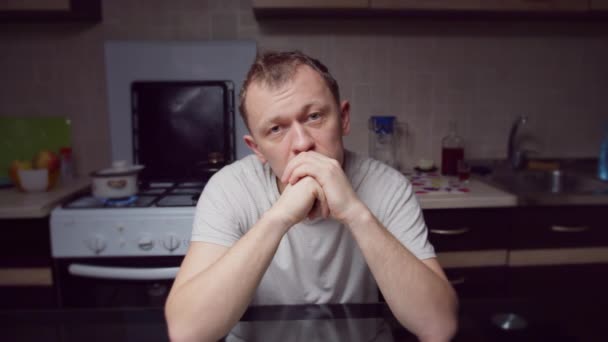 Um homem deprimido está sentado à mesa da cozinha à noite, olhando diretamente para a câmera, movimento da câmera — Vídeo de Stock