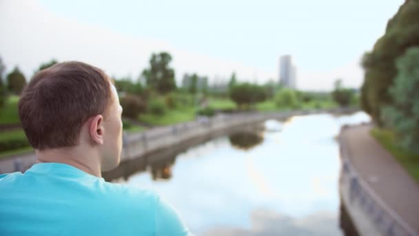 파란색 티셔츠를 입은 한 남자가 다리 위의 공원에 서서 강을 바라보고 있습니다. 카메라의 움직임, 복사 공간, 클로즈업 — 비디오