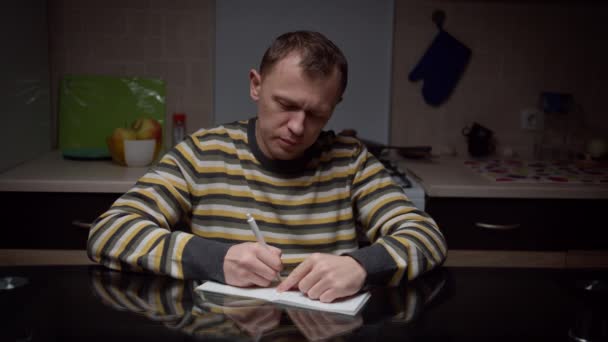 El hombre escribe en un cuaderno sus planes, sentado en la mesa de la cocina por la noche — Vídeos de Stock