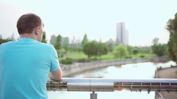 Man staat in een park op een brug en kijkt naar de rivier, camerabeweging, kopieerruimte — Stockvideo