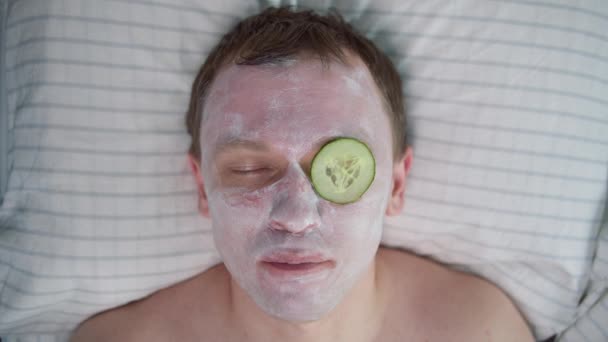 男は顔の世話をし 顔に化粧マスクをして横になり キュウリのスライスを食べる — ストック動画