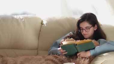 Gözlüklü esmer kız kanepeye oturmuş kitap okuyor, sayfaları çeviriyor, fotokopi çekiyor, kamera çekiyor.