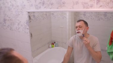 Adam sabah sakalını kesiyor. Banyoda aynanın karşısında dikiliyor. Kamera hareket ediyor.