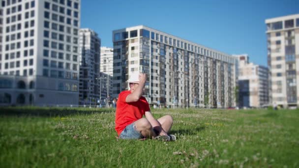 一个戴帽子的男人坐在公园的草地上 背景是城市的风景 镜头在移动 — 图库视频影像