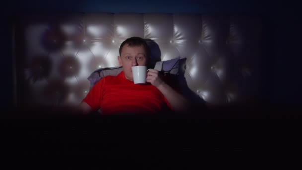 一个年轻人对他在电视上看到的非常惊讶 他晚上躺在床上 手里拿着杯子 镜头在动 — 图库视频影像