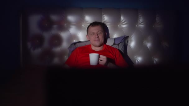 年轻迷人的小伙子喝了一大杯茶 正在晚上看电视 看镜头 — 图库视频影像